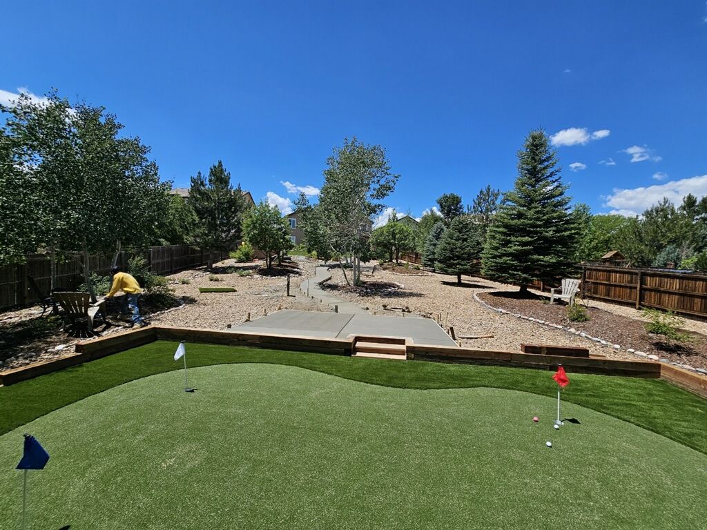 Backyard putting green with flags, trees, and gravel landscaping under a blue sky.