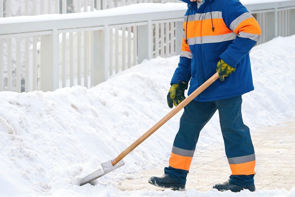 Person in blue and orange work clothes shoveling snow on a sidewalk, near a white fence.
