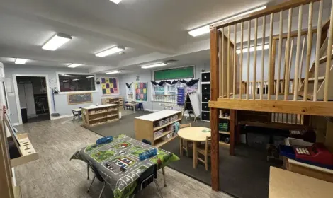 A wide-angle view of a colorful, organized preschool classroom with a wooden indoor play loft, tables, and toys.