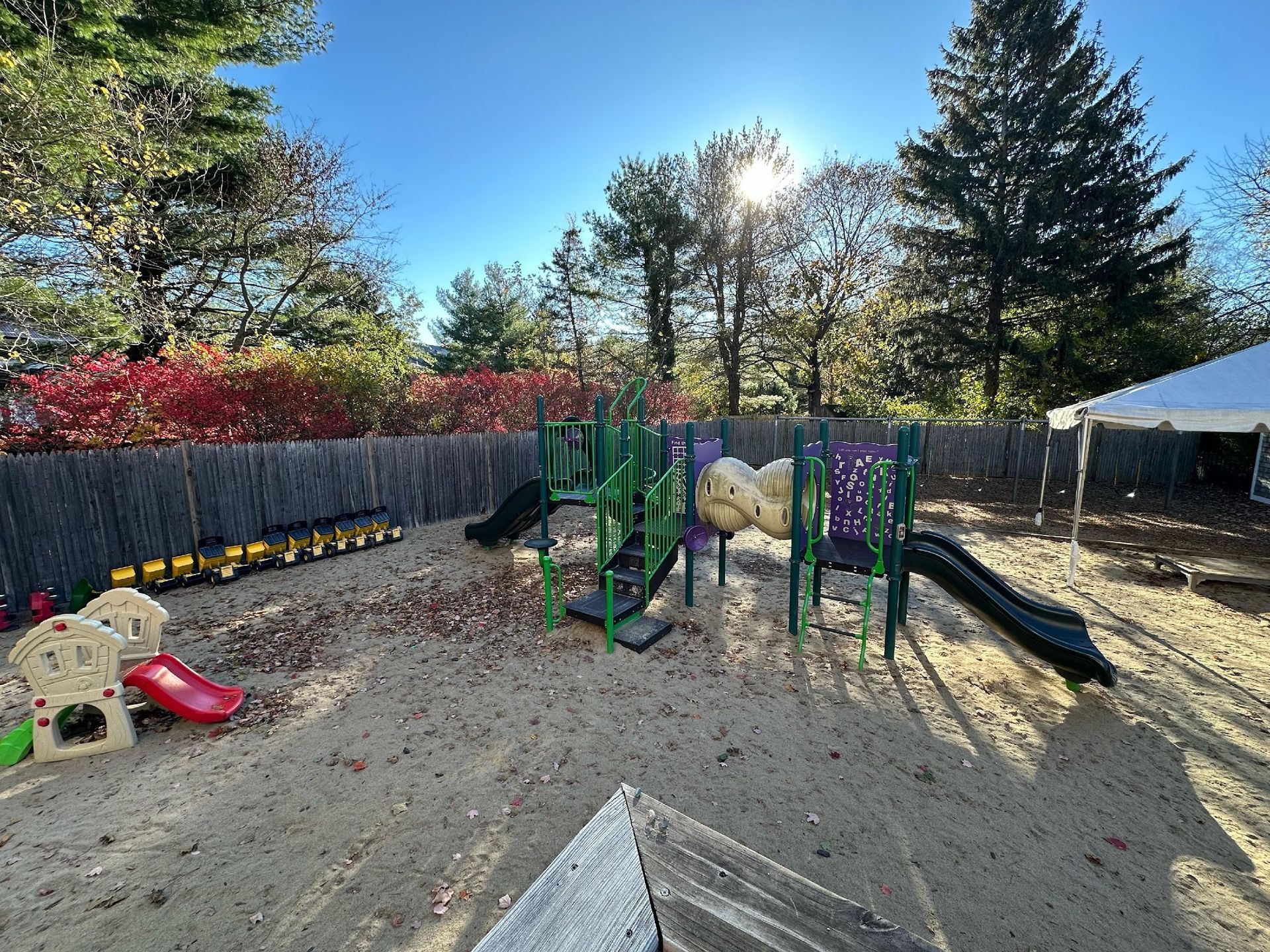 A playground with a green climbing structure, slides, and a small play set on a sandy lot against a tree-lined backdrop.
