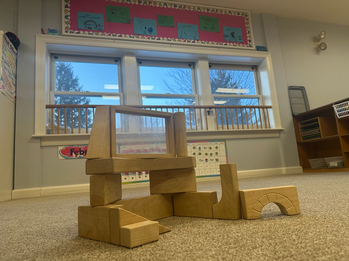 A wooden block structure built on a carpeted floor in a classroom, with large windows and a bulletin board in the back.