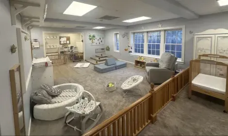 A bright infant childcare room with a crib, play mats, bouncers, and a soft-toned sitting area with carpeted floors.