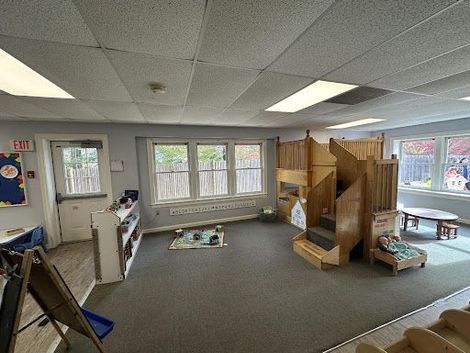 A child's playroom features a large wooden play structure, a play mat, shelving, and a small table on a gray carpet.
