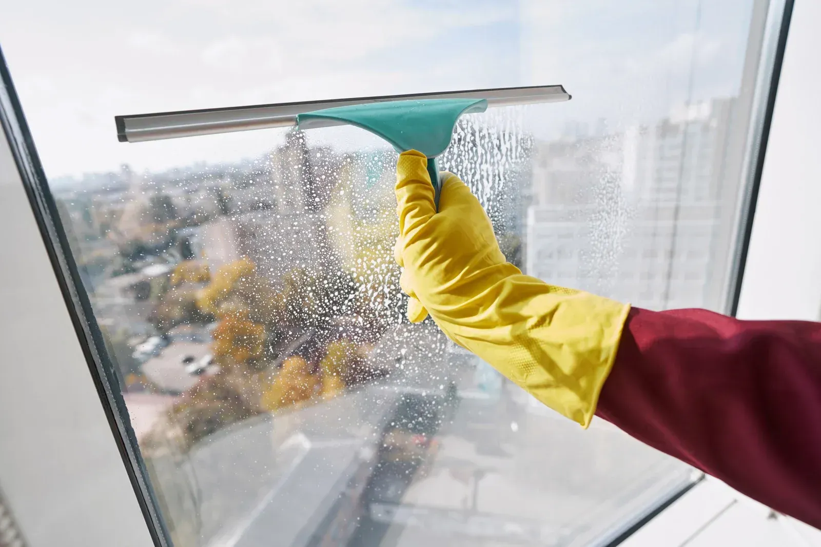 Person in yellow glove uses a squeegee to clean a window. City buildings in the background.