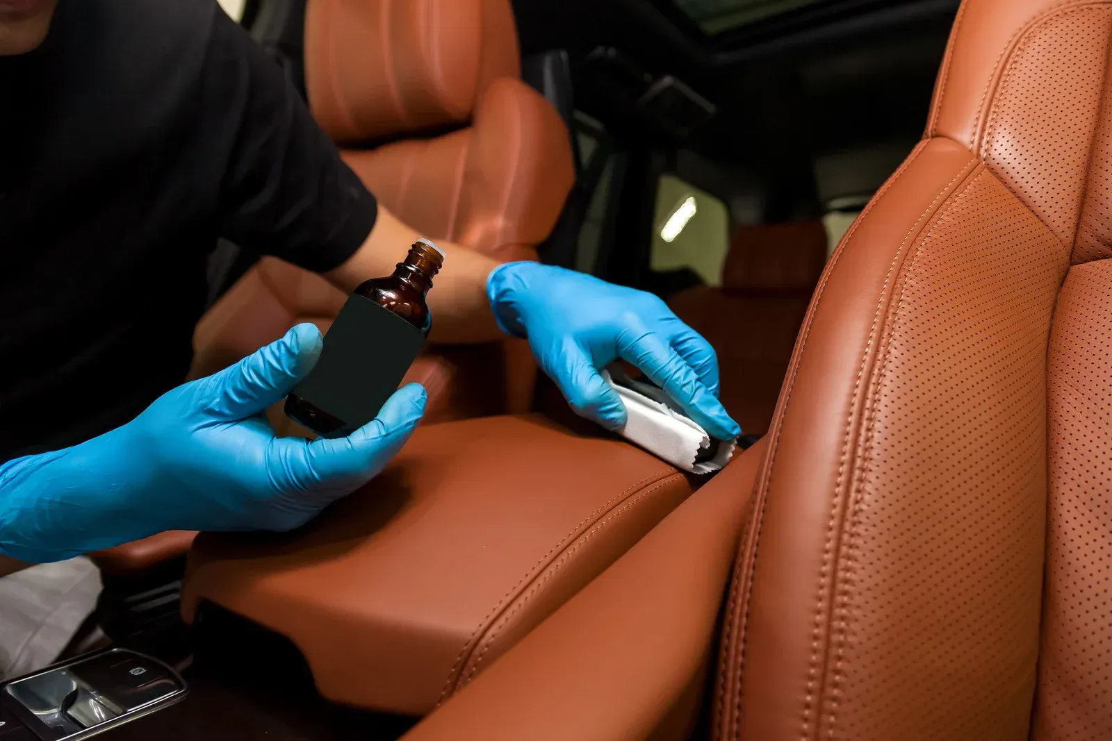 Hands in blue gloves applying leather protectant to a brown car seat with a bottle and applicator.