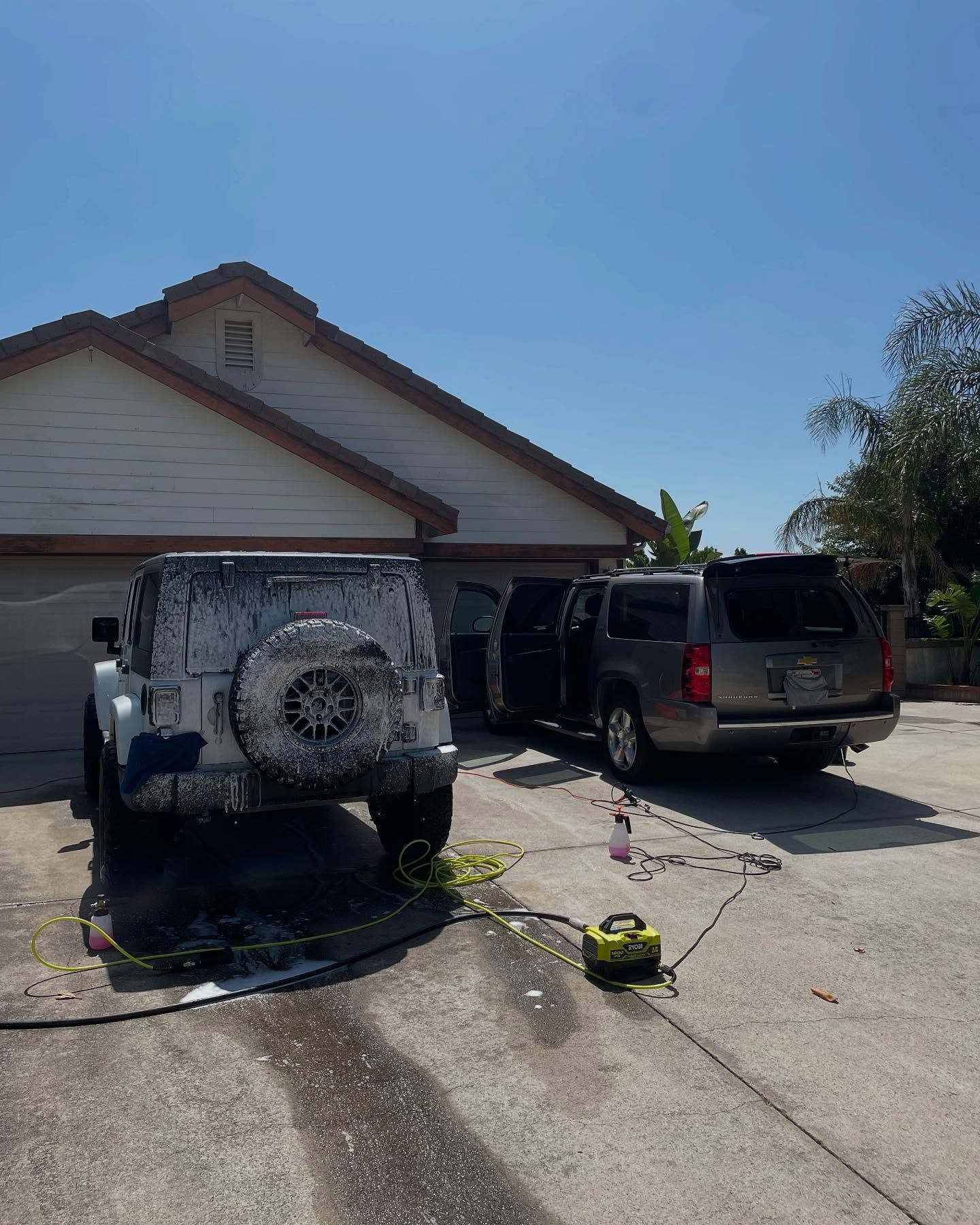 Cars being washed in a driveway on a sunny day. A Jeep covered in foam sits next to a SUV with open doors.