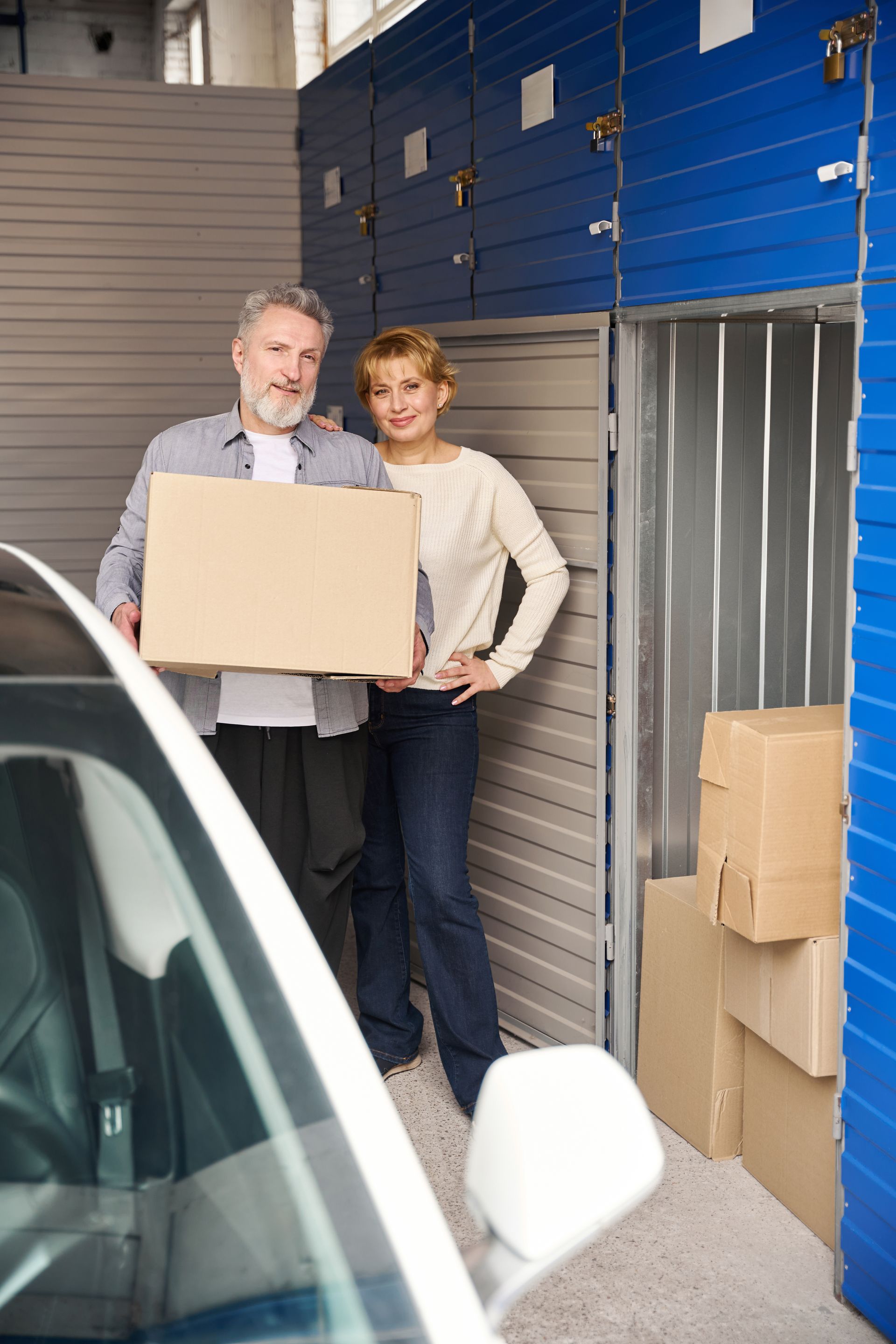 Smiling couple holding a cardboard box next to their car beside an open storage unit full of boxes.