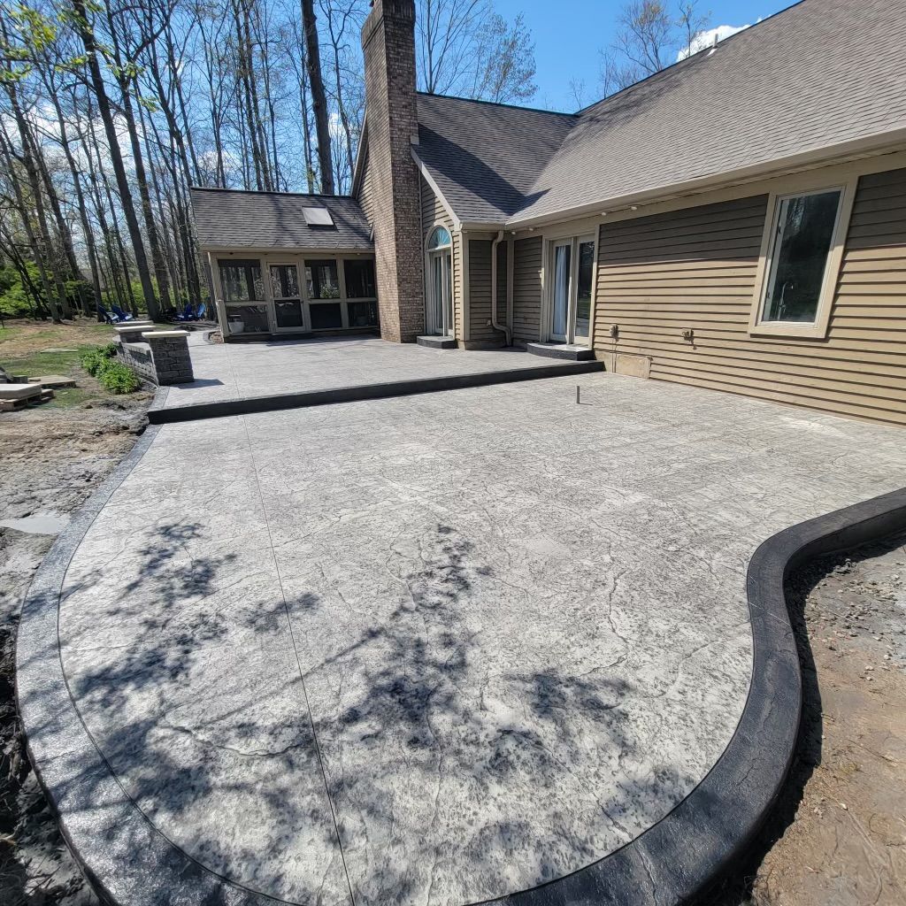 New concrete patio with a curved edge next to a house with a chimney and surrounding trees.