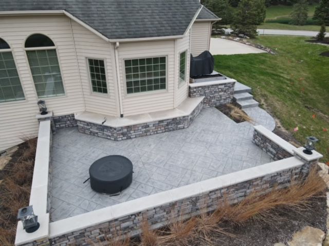 Stone patio with retaining walls next to a house with steps leading to grassy area.