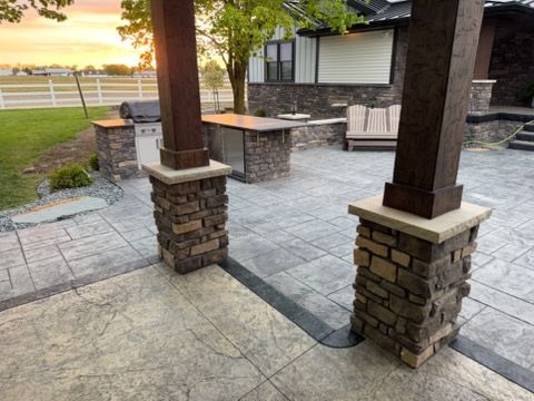Covered stone patio with columns, paver flooring, and outdoor seating beside a grassy field at sunset