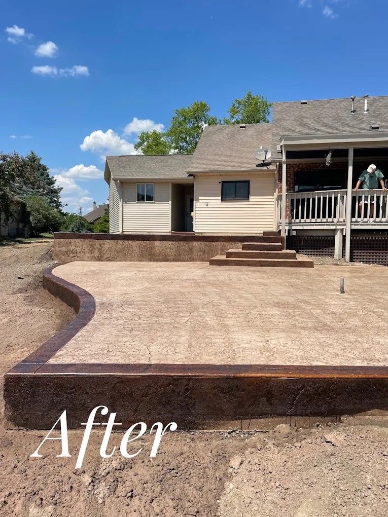 Backyard concrete patio with curved border and steps, beside a beige house under a blue sky.