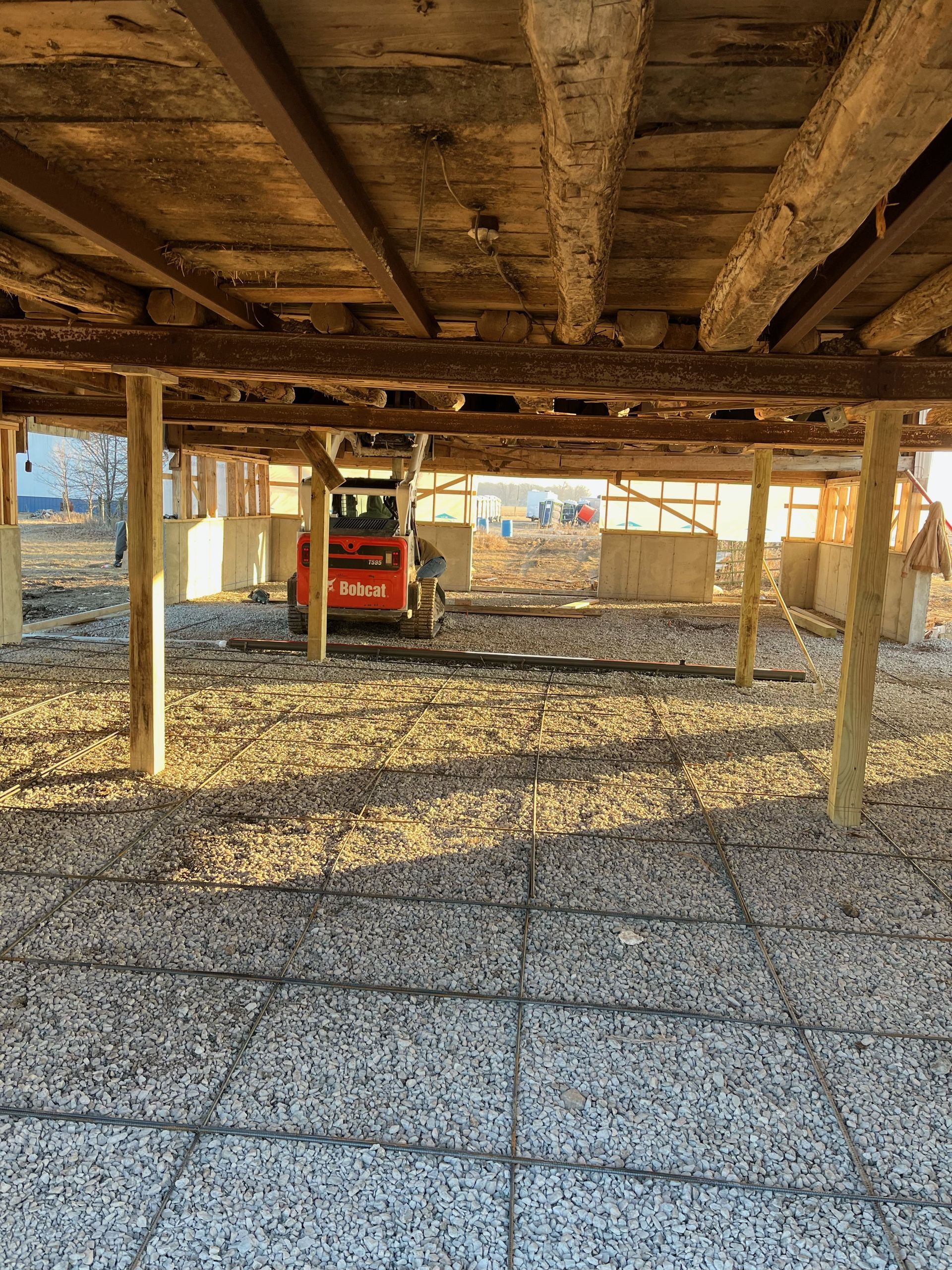 View from under a raised wooden structure, supported by posts, over a stone-covered ground with construction equipment in the background.