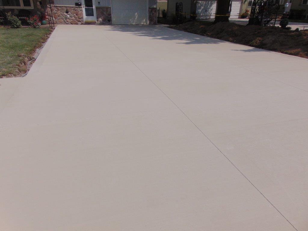 Light tan driveway with cracks. A house is visible in the background with a garage.