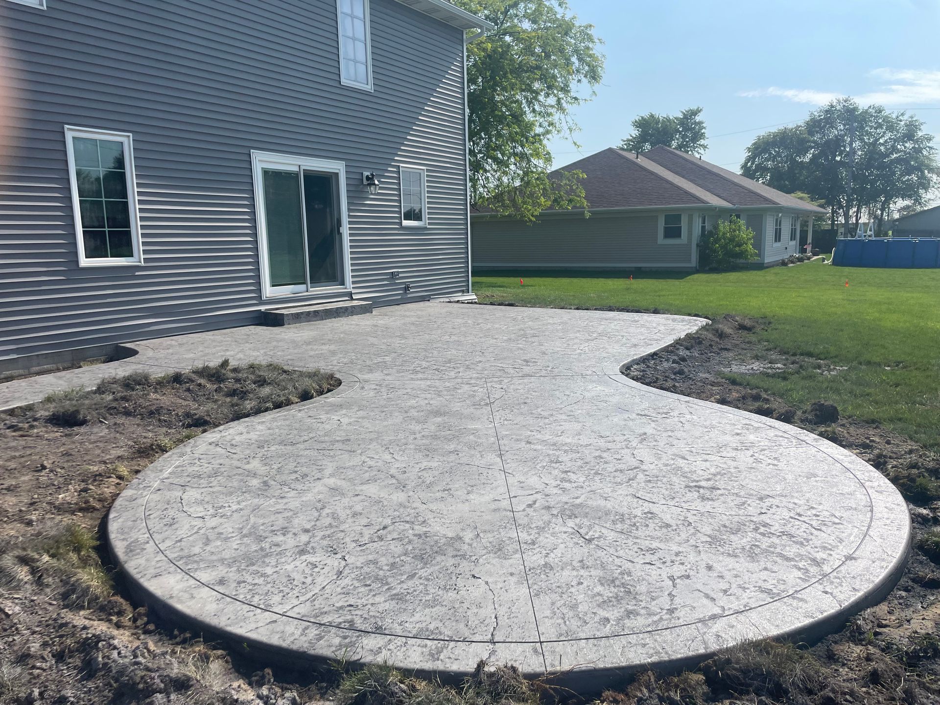 Backyard patio under construction beside a gray house, with a curved gravel base and lawn nearby