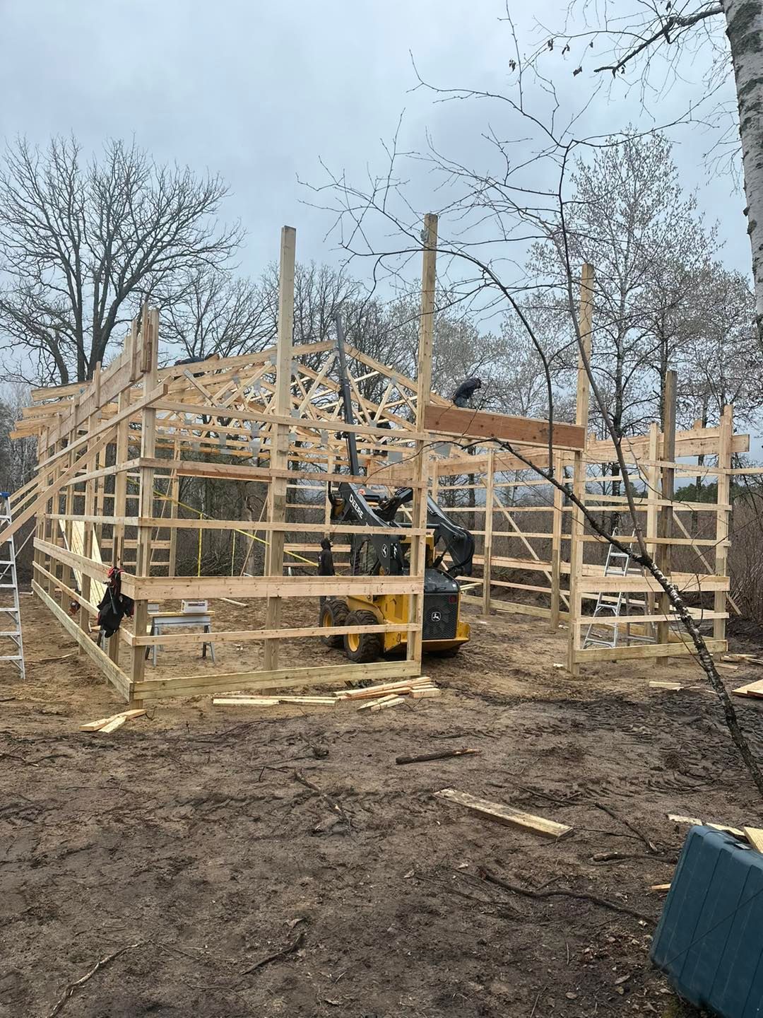 Partially framed wooden building under construction on a muddy lot with bare trees in the background