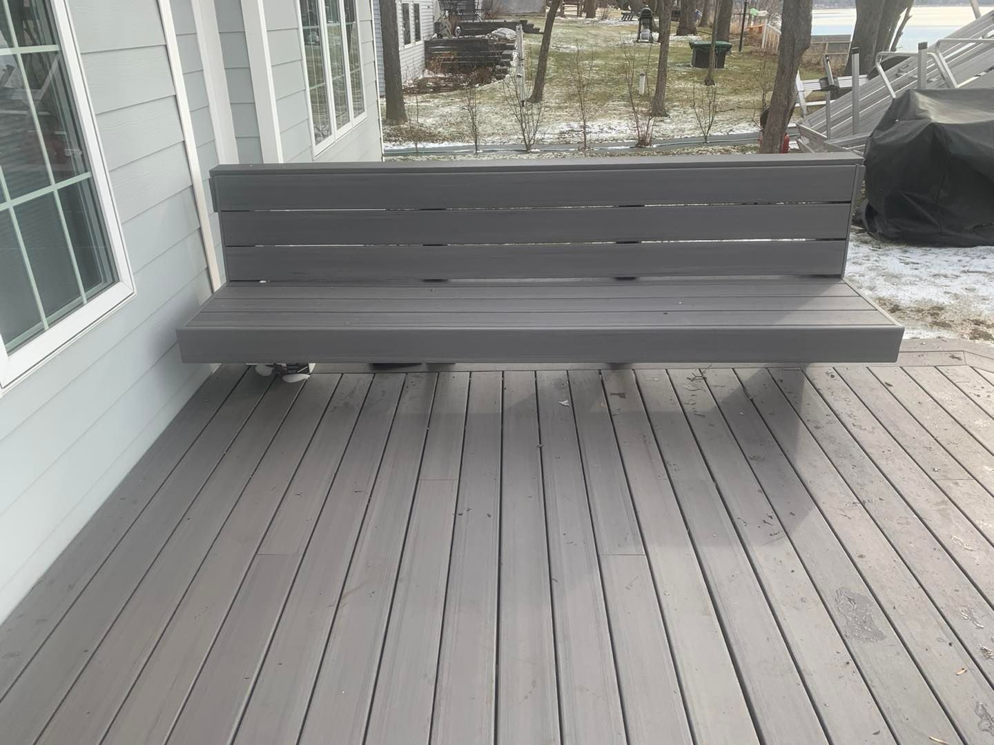 Gray bench on a wooden deck beside a white house, with a backyard and trees in the background