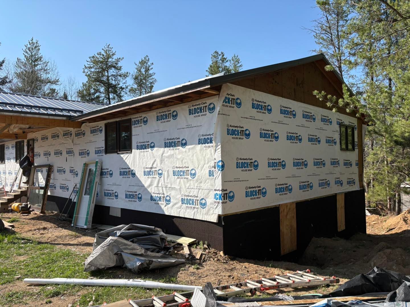 House under construction wrapped in white housewrap, raised on stilts in a wooded area