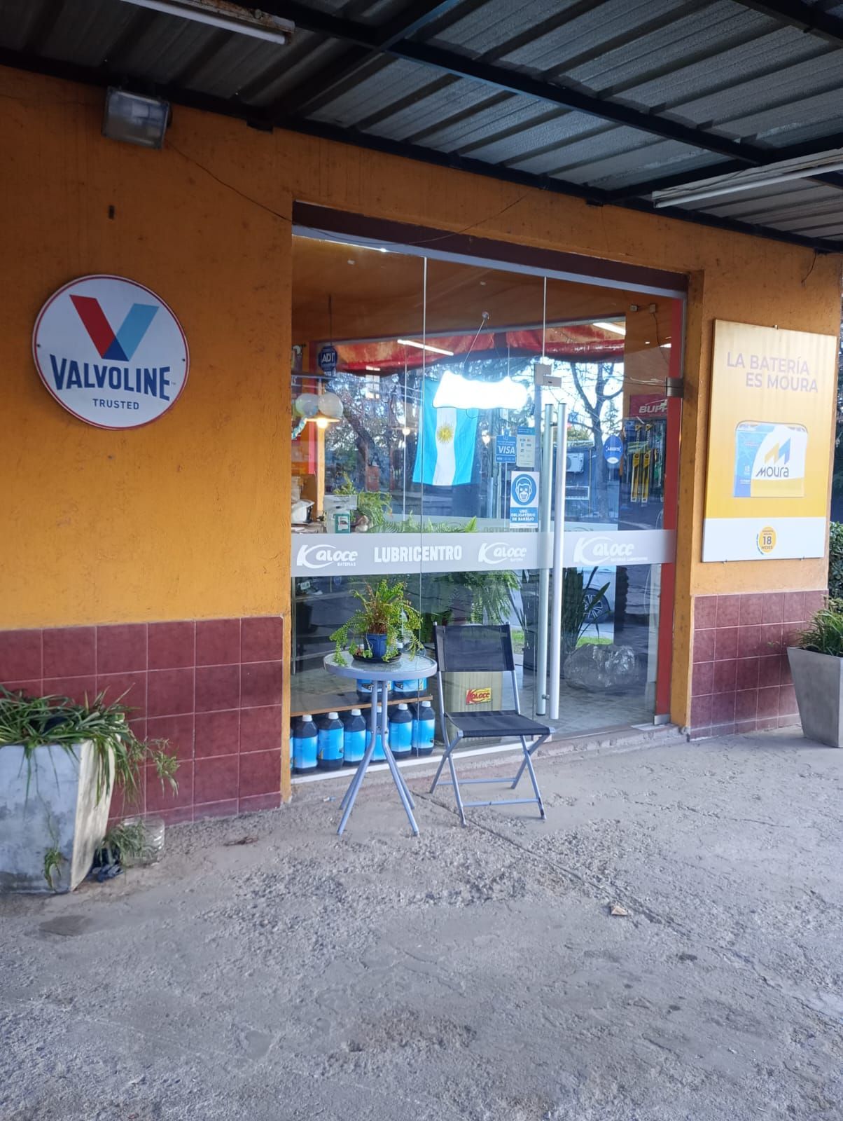 Pequeña tienda con paredes naranjas, puerta de cristal y una silla bajo un toldo metálico.