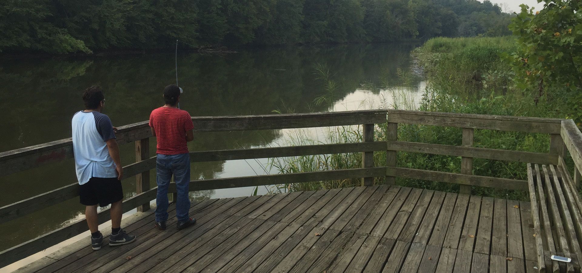 Two people standing on a wooden deck overlooking a calm river and green trees
