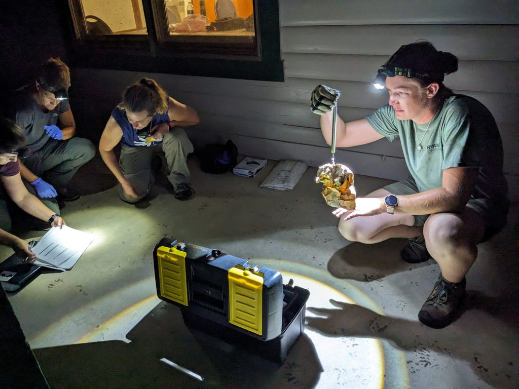 Three people crouch in a dark room, examining a device with flashlights and a bright work light.