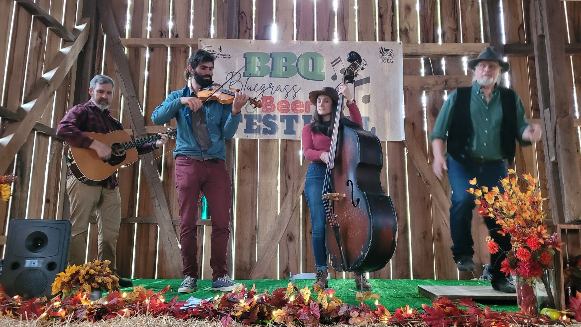 Four musicians perform on a decorated stage with banner backdrop and autumn floral arrangements.