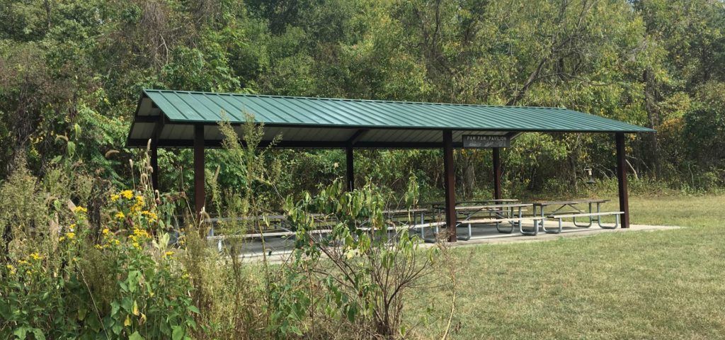 Open-sided green-roof pavilion in a grassy park with picnic tables, trees behind it