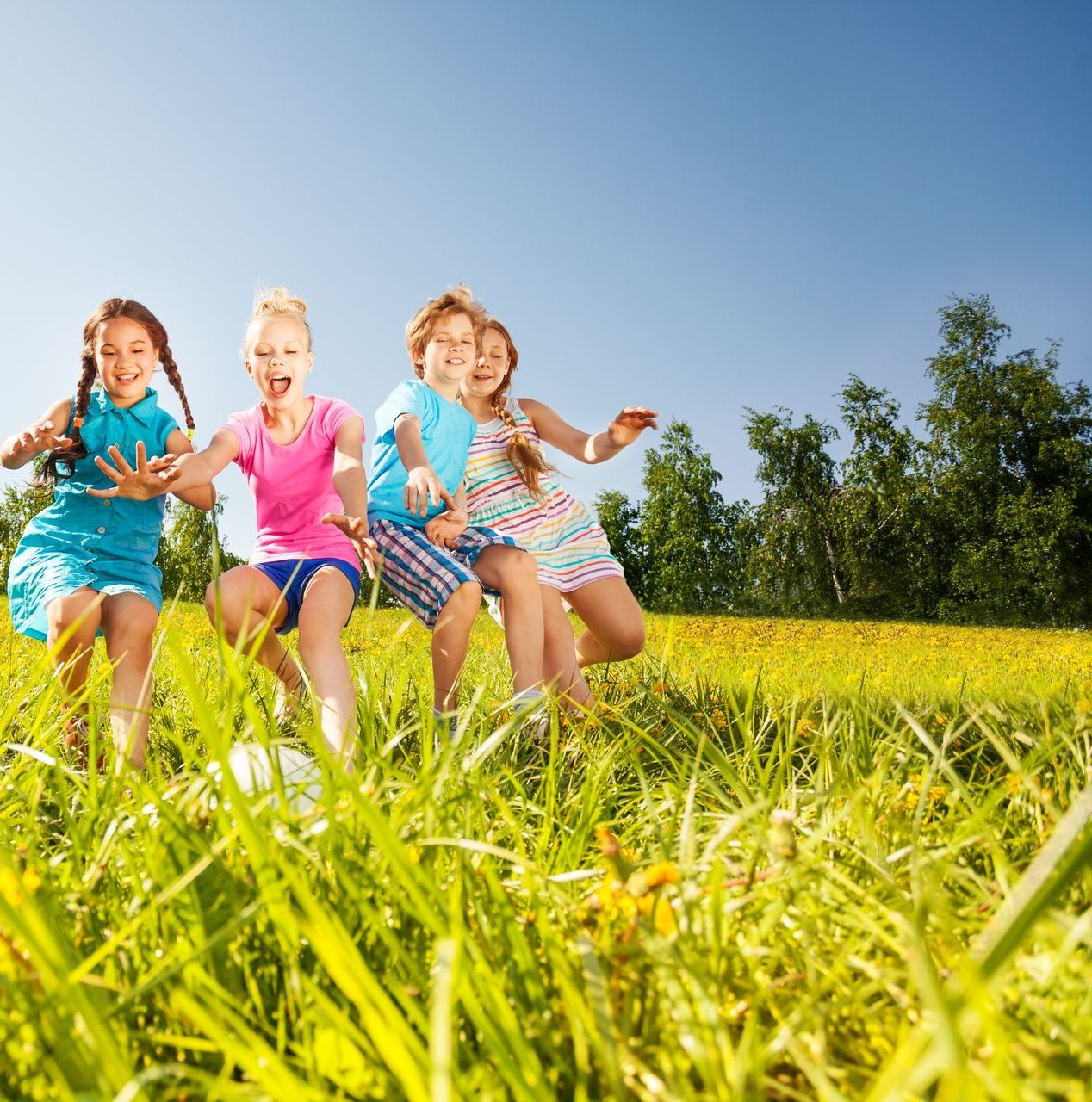Several children in colorful clothing are running through the grass with trees in the background.