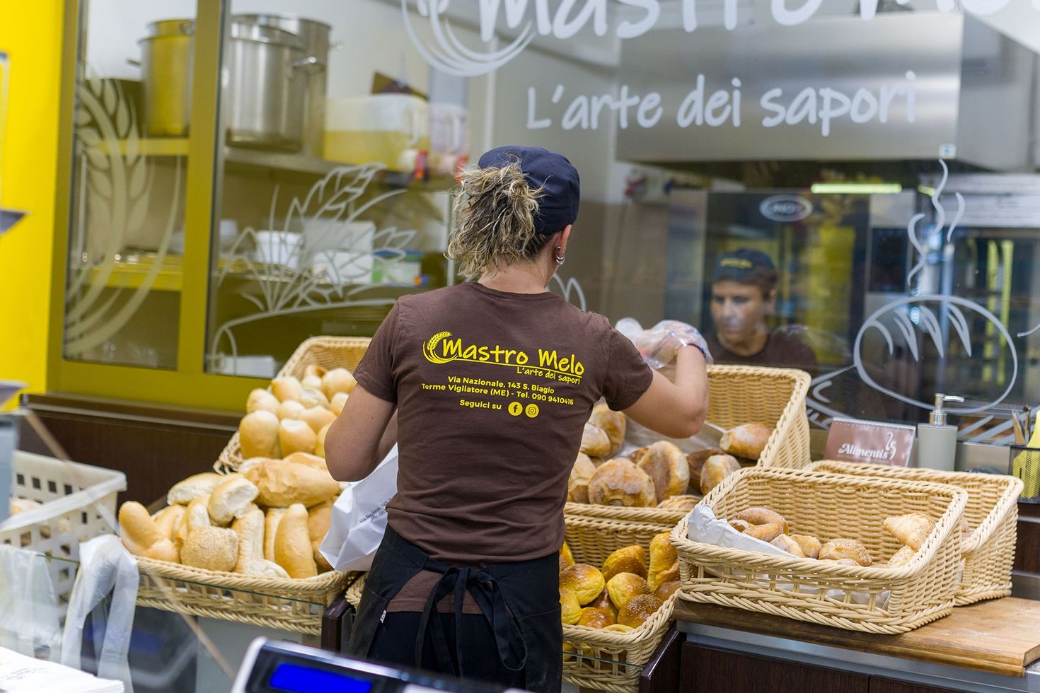 Bancone di panetteria con cesti di pane e un addetto che sistema le pagnotte dietro la vetrina.