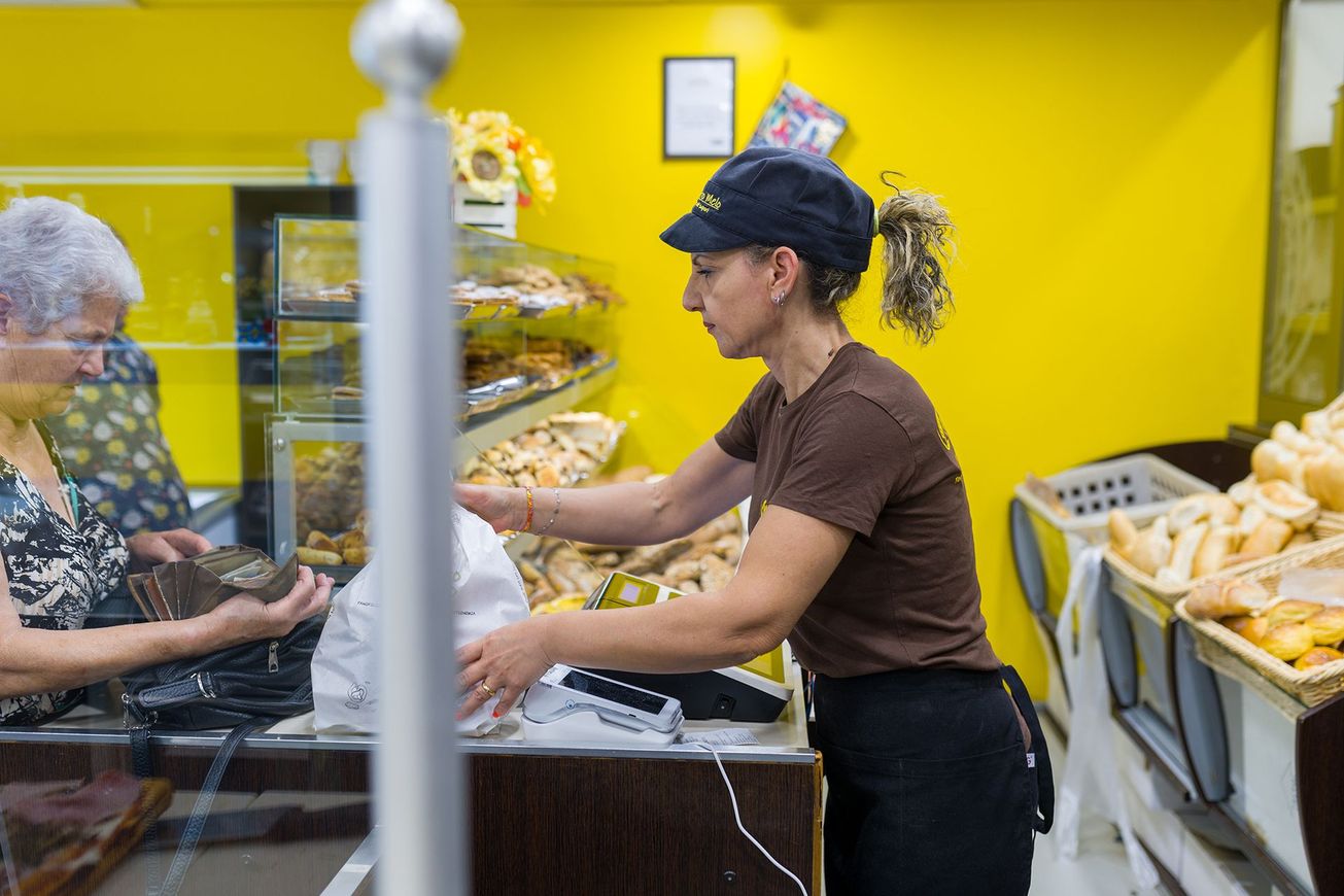 Bancone di panetteria con una cassiera che porge i prodotti a un cliente in un negozio giallo brillante.