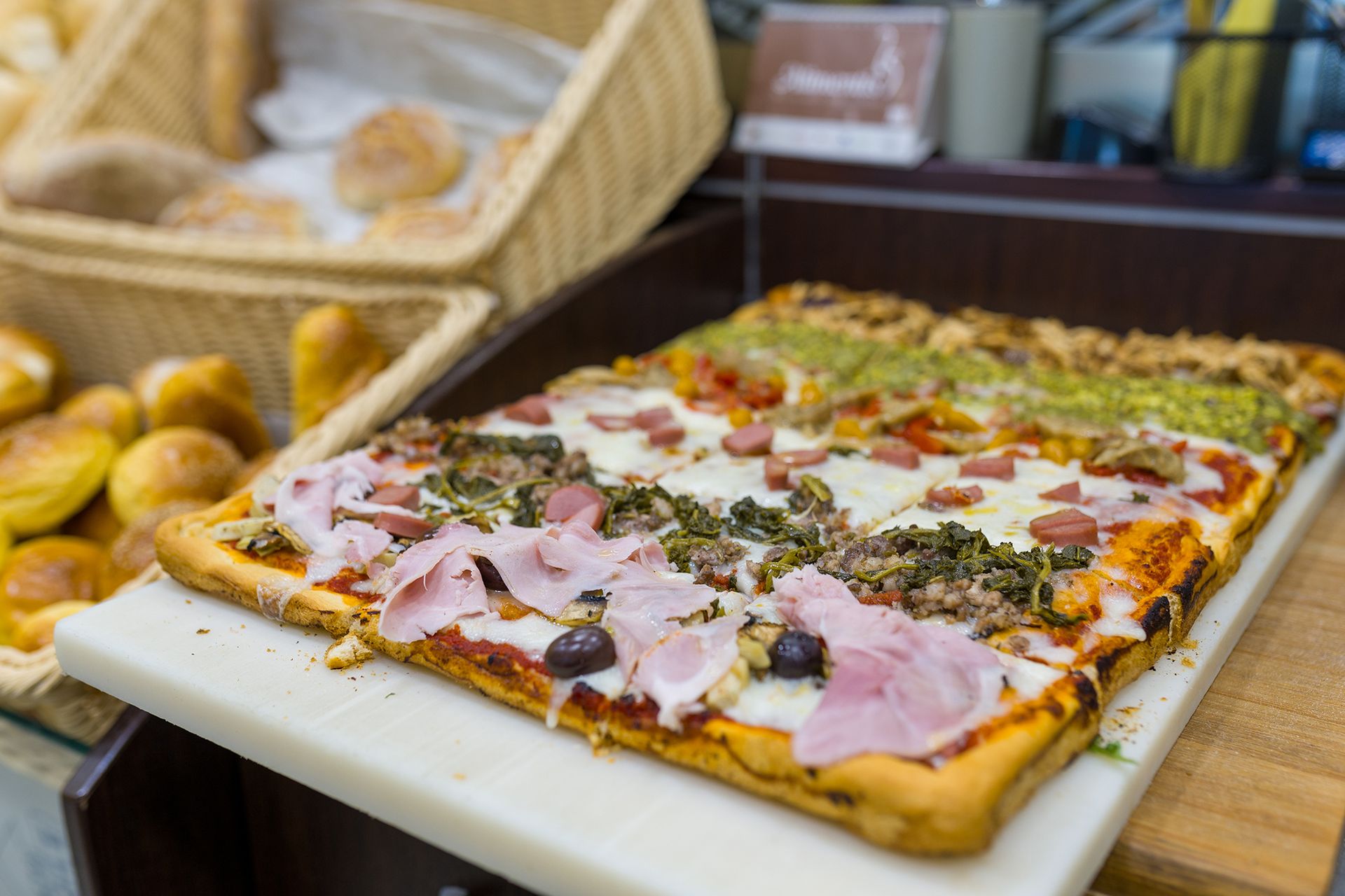 Pizza rettangolare con prosciutto, olive e verdure su un bancone in una panetteria