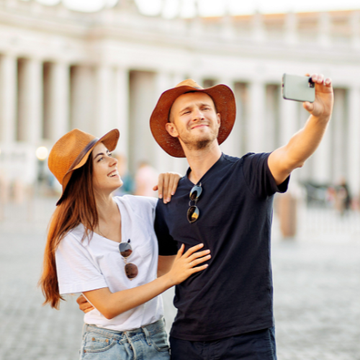 Two tourists in hats take a selfie in front of a bright building, smiling outdoors.