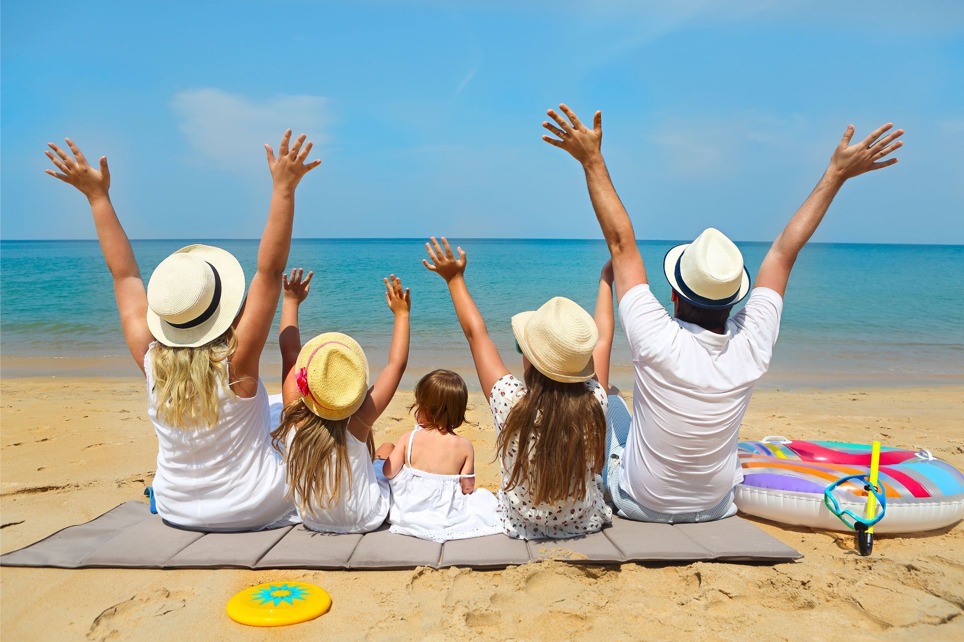 Four people sit on a beach with arms raised toward the ocean, wearing sun hats beside colorful beach toys.