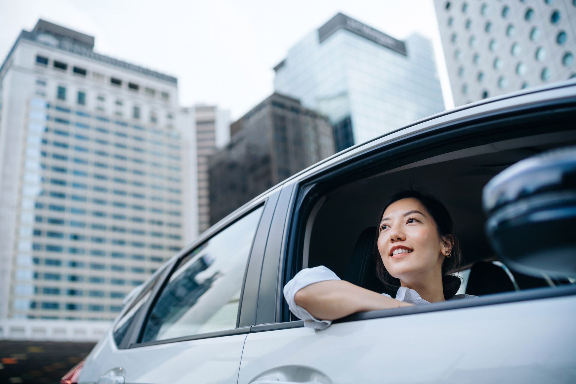 Woman smiles out car window in a city with tall buildings.