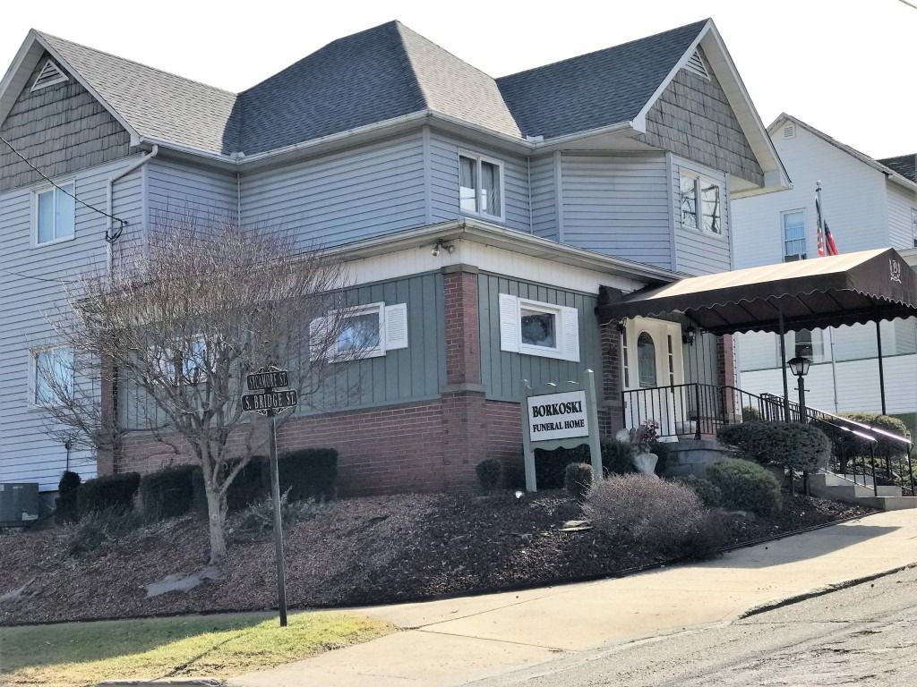 Two-story gray-and-white building with a front porch and sign, on a sunny street slope