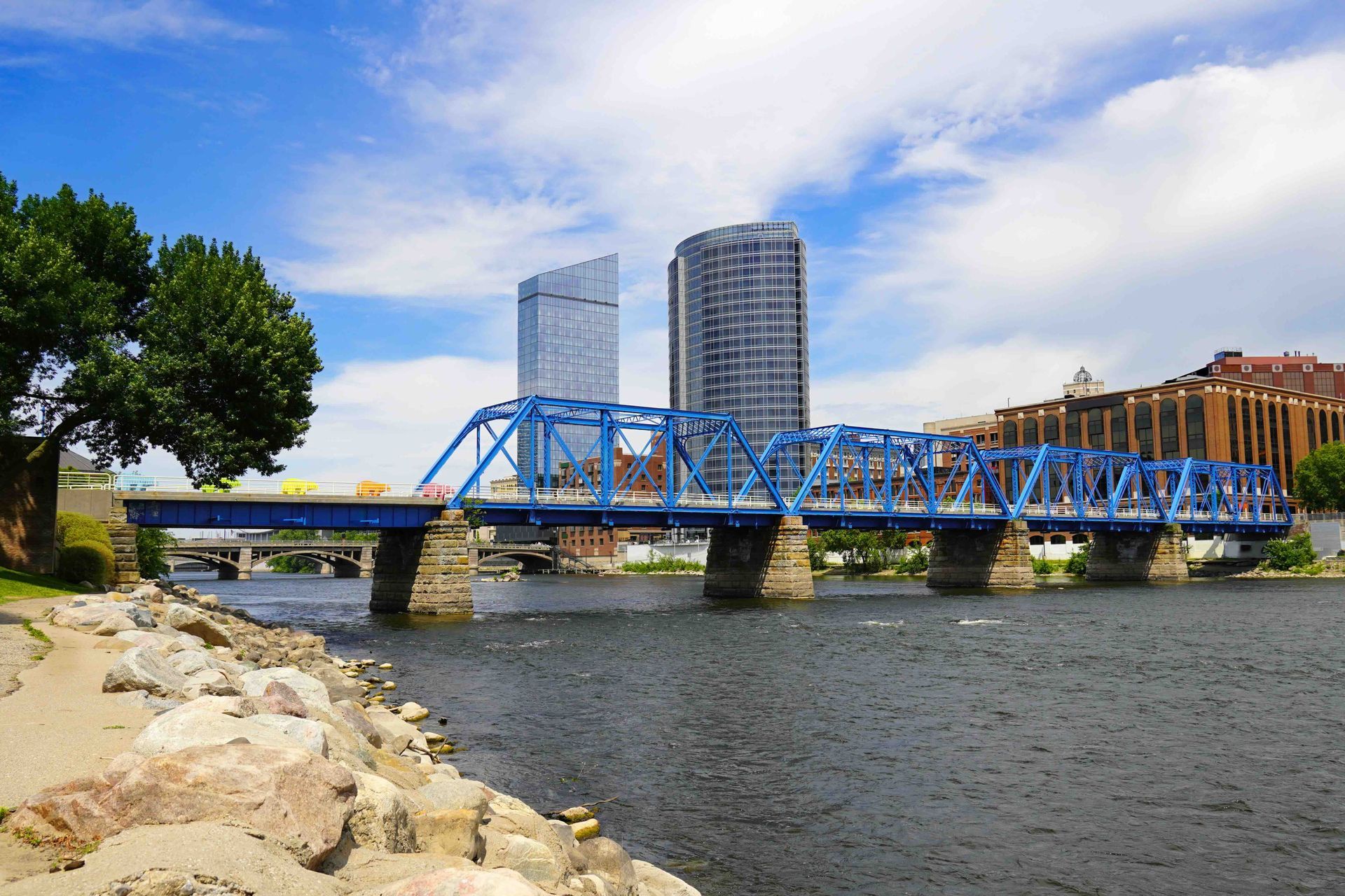 Blue steel bridge over a river with city buildings and rocks in the foreground under a partly cloudy sky