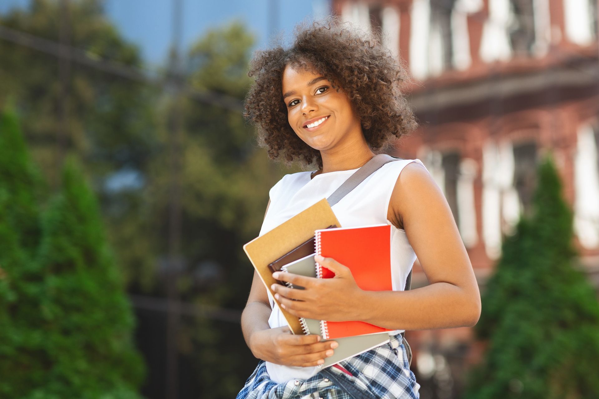 Student smiling outdoors, holding books and a red notebook in front of a campus building