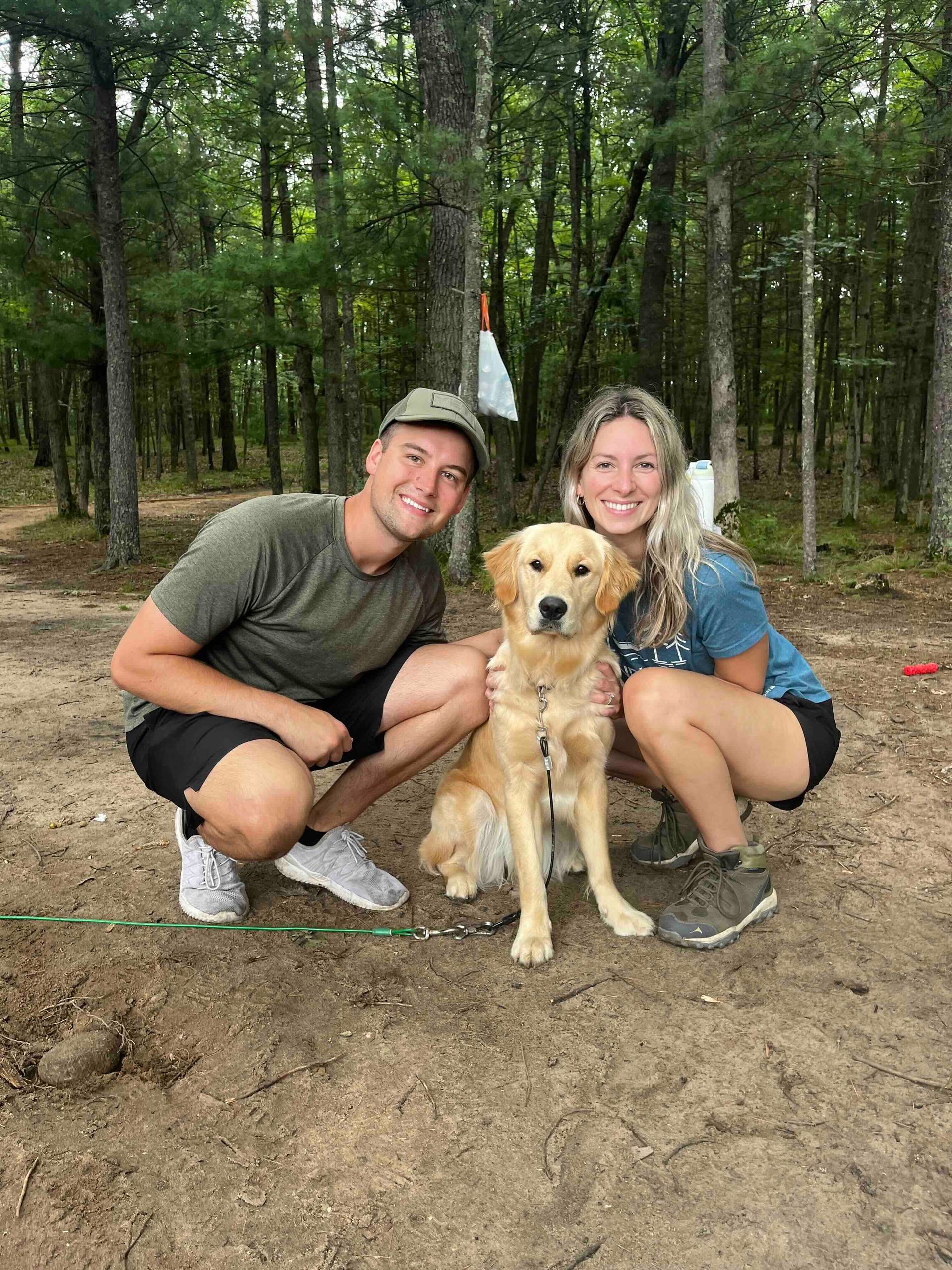 A man and a woman are squatting down next to a dog in the woods.