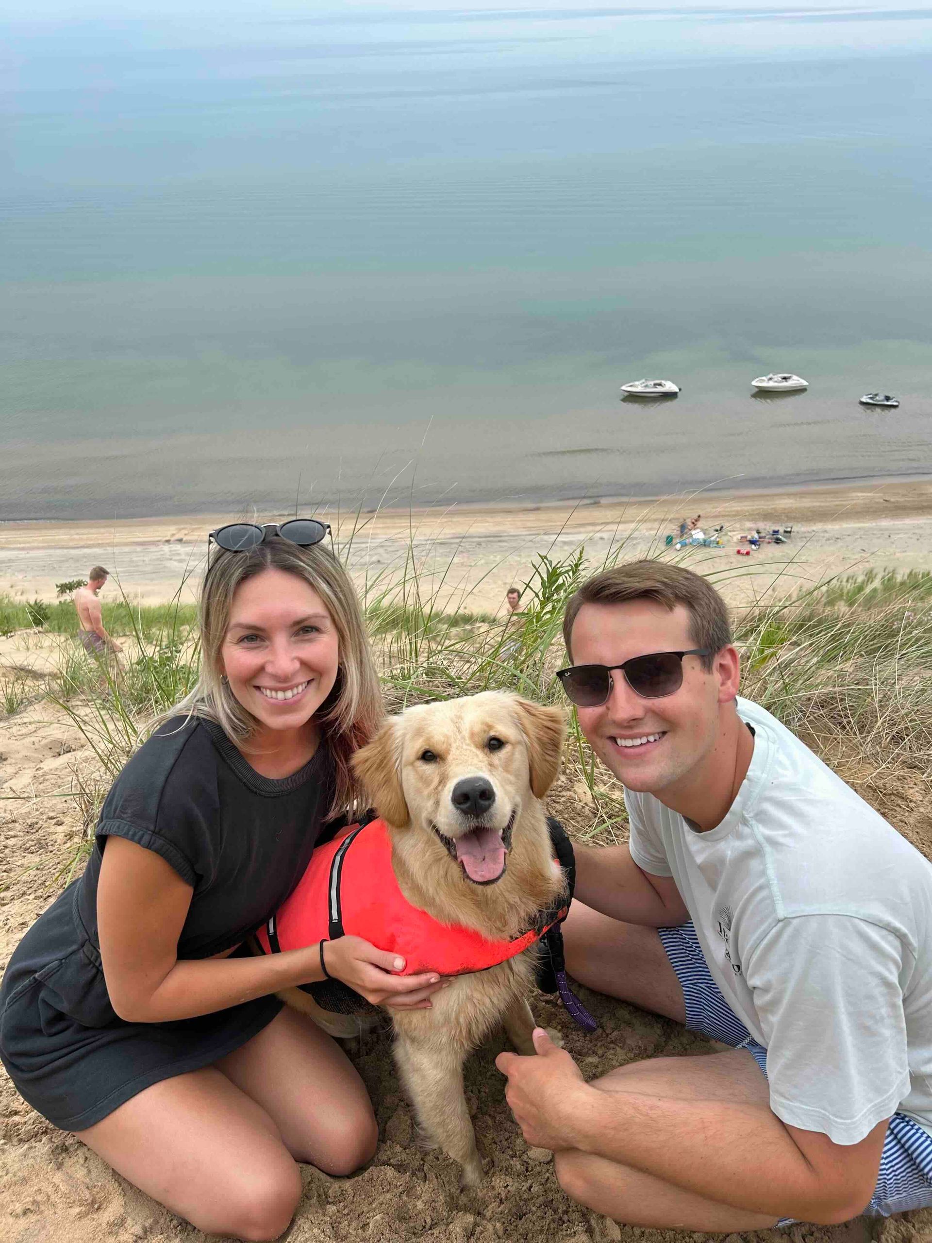 A man and a woman are kneeling down next to a dog on a beach.