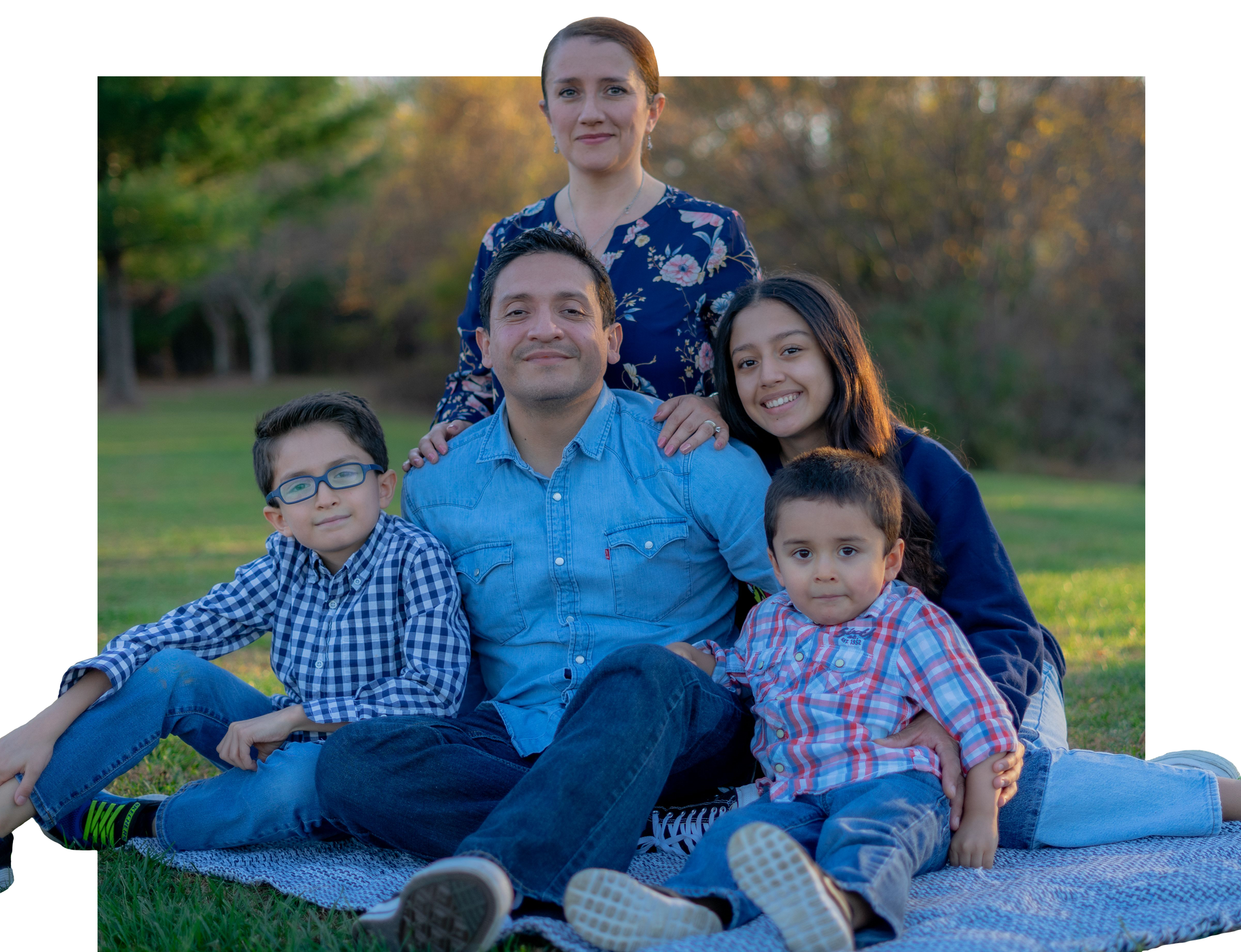 Family sitting on a blanket outdoors, smiling in a park with trees and grass.