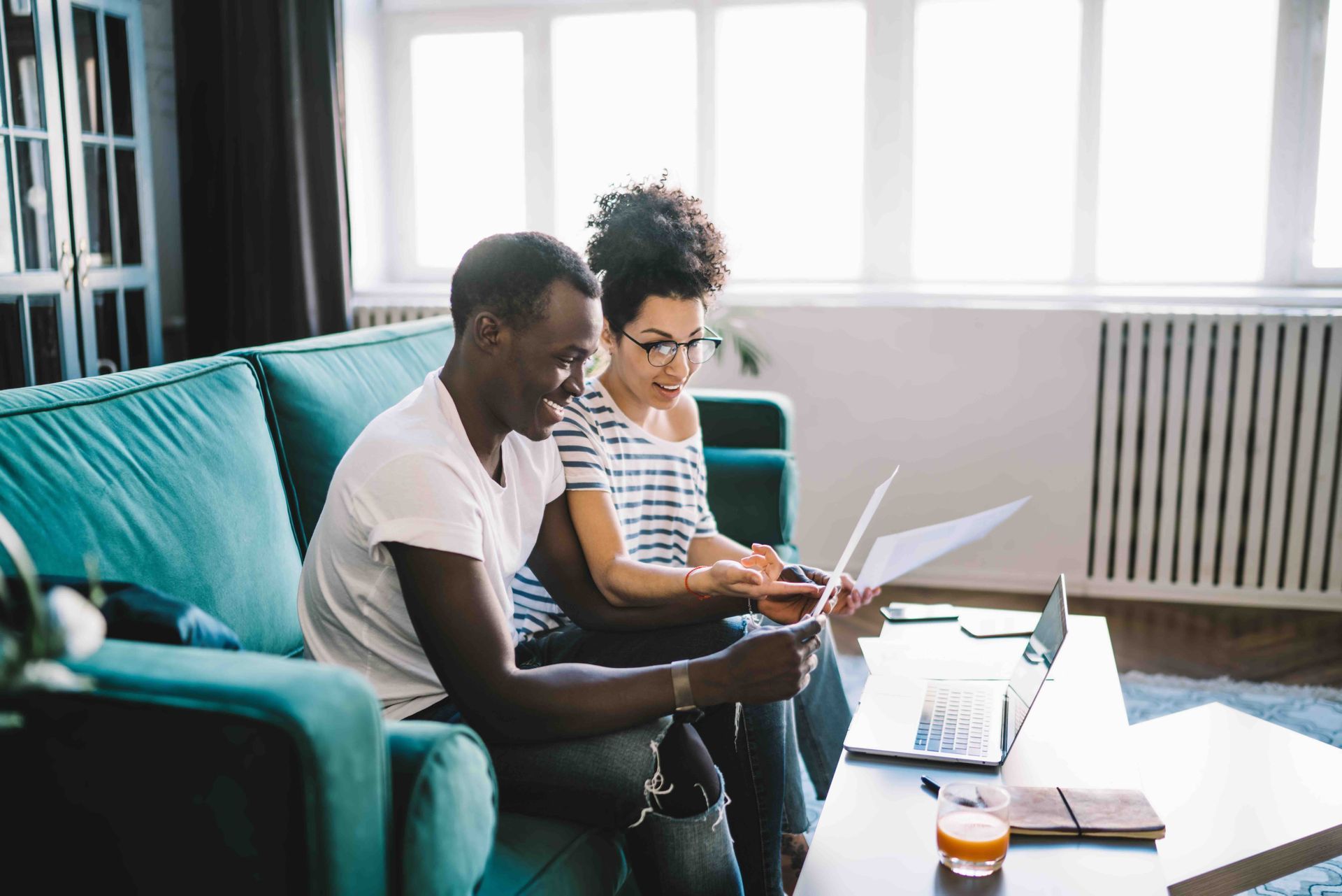 Couple on a teal couch, reviewing documents and using a laptop in a well-lit living room.