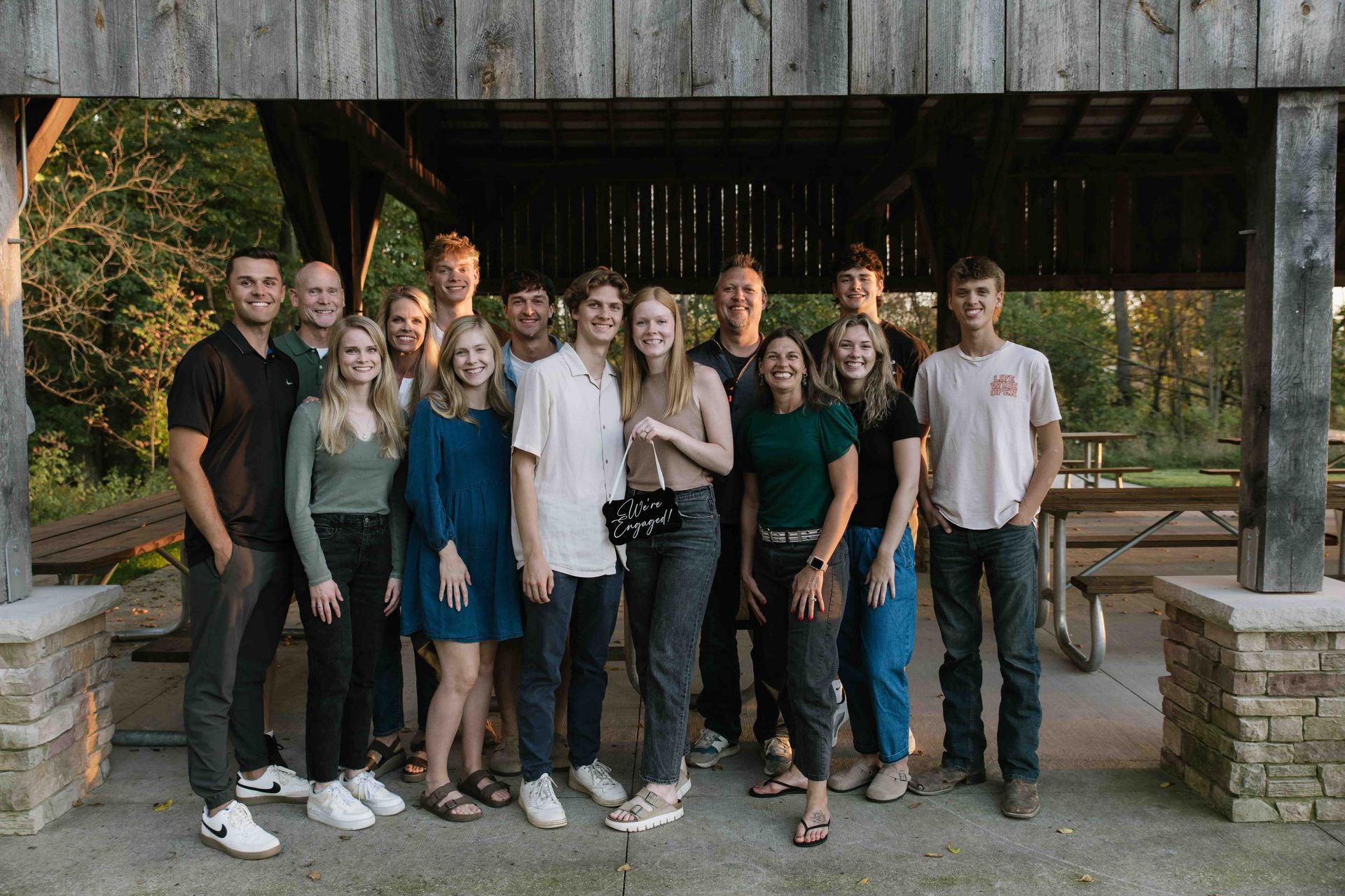 Group of people smiling under a wooden shelter, outside. Daylight casts warm tones.