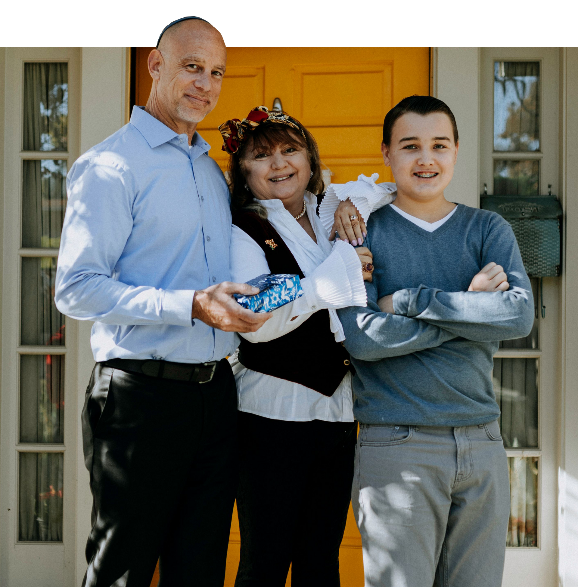 Three people standing on a porch, holding wrapped items and smiling in front of a yellow door.