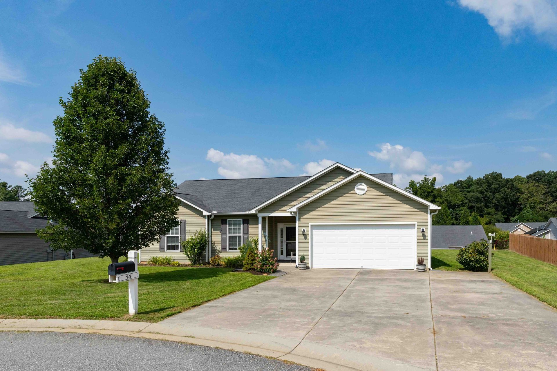 Green house with white garage door, driveway, green lawn, tree, blue sky.