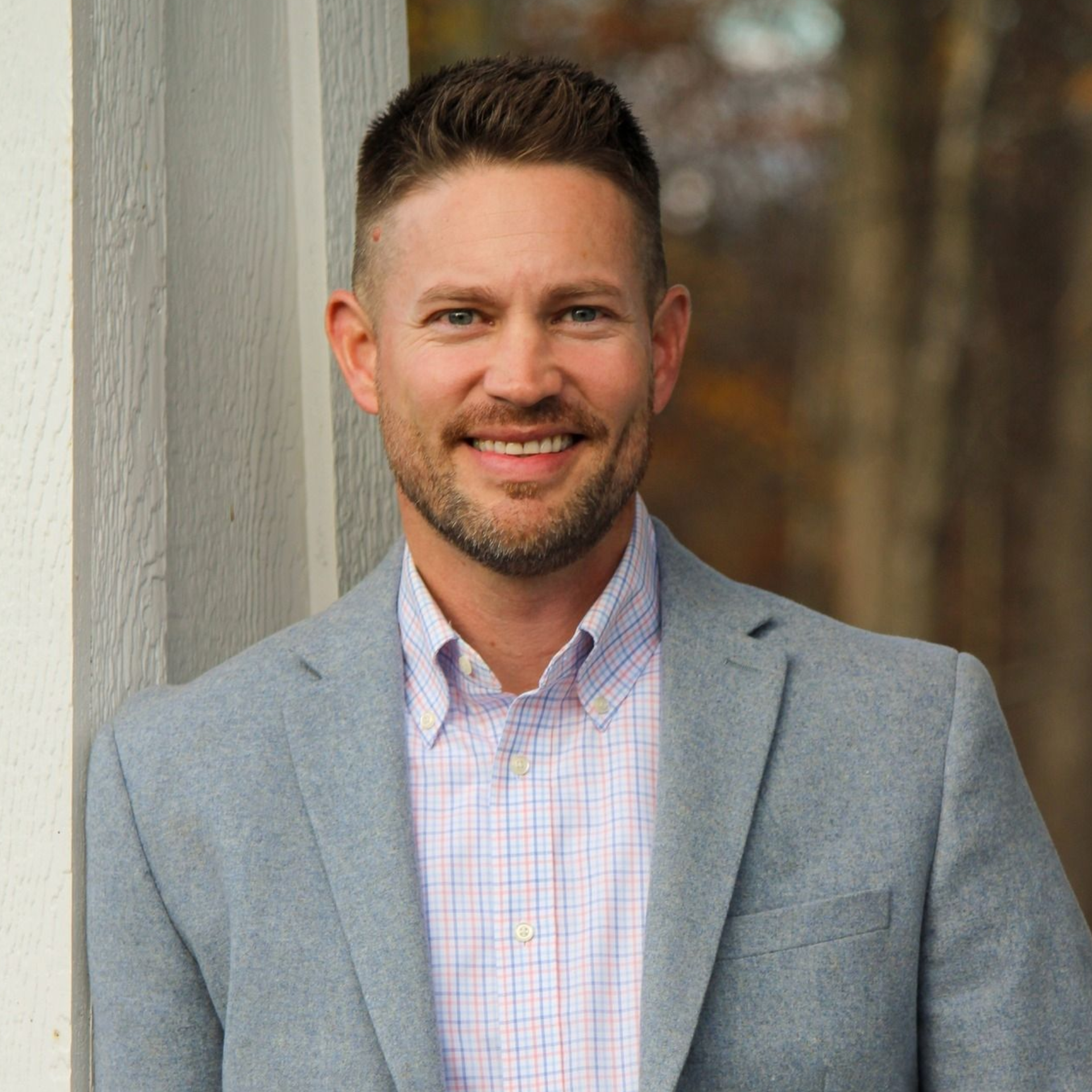Smiling man in a gray blazer and checkered shirt standing by a wooden wall outdoors