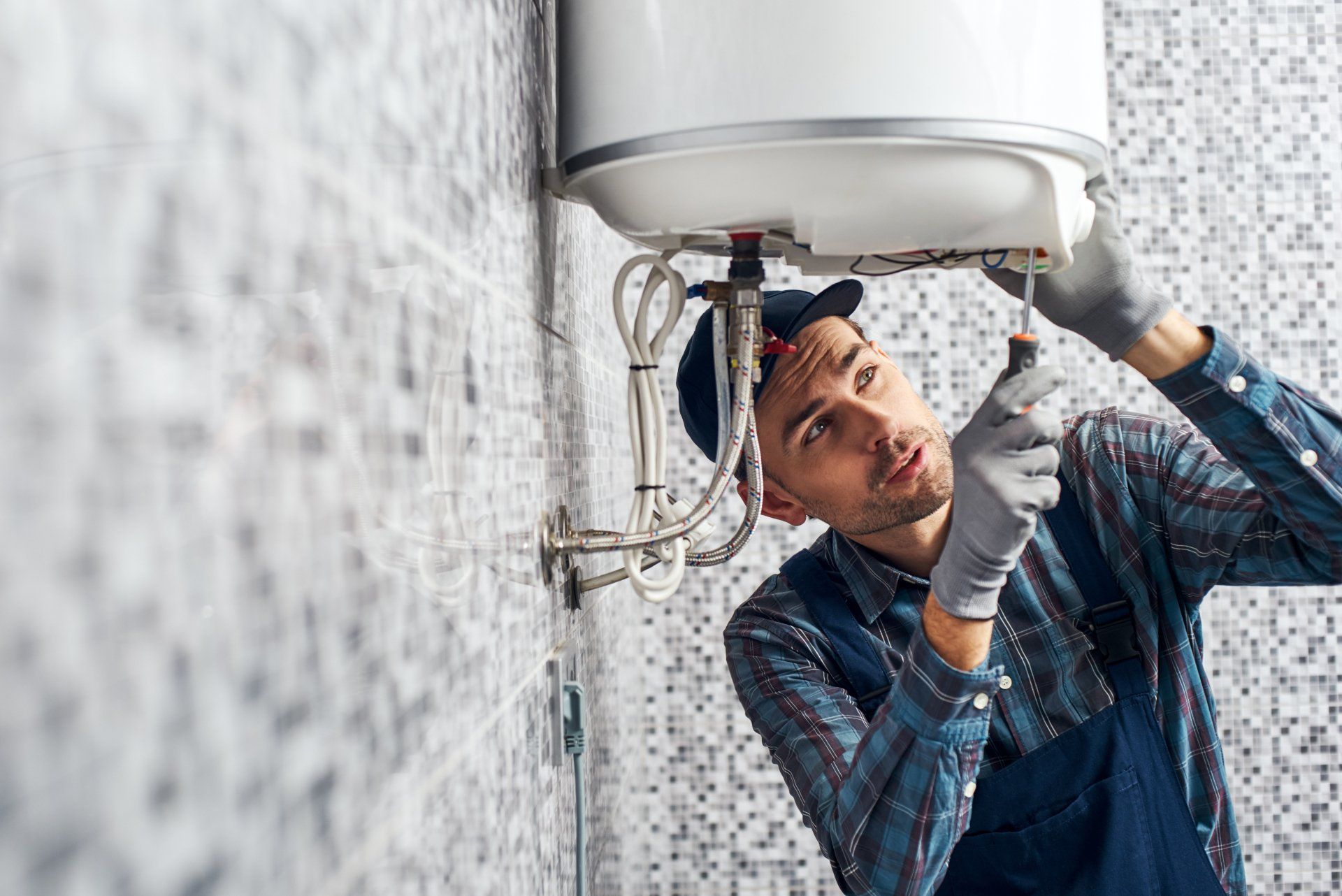 Technician repairing a wall-mounted water heater, wearing gloves and tools, in a tiled room