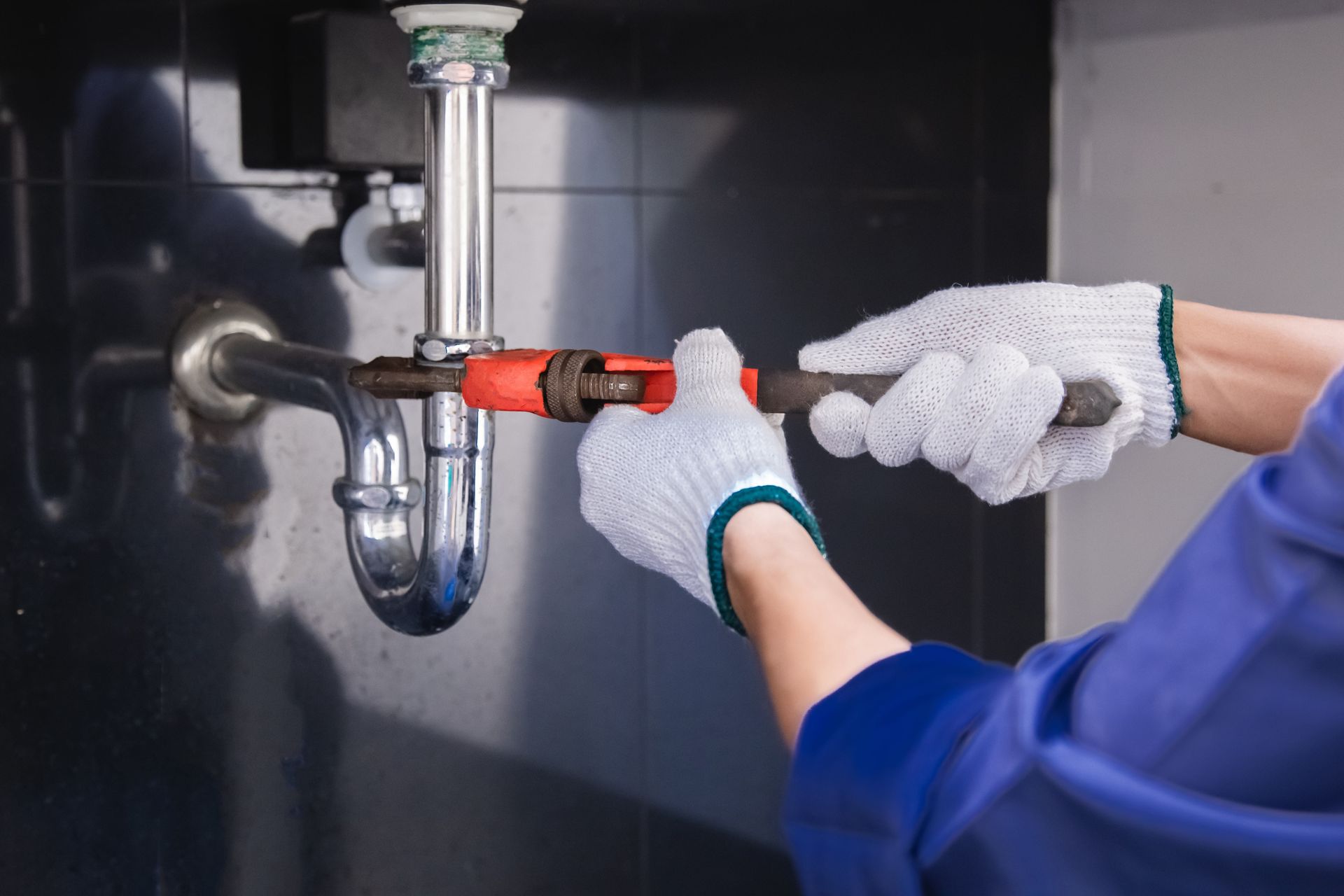 Hands in gloves tightening a pipe under a sink with a red wrench tool