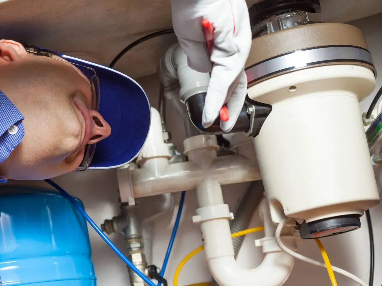 Technician inspecting under-sink plumbing with flashlight, beside a blue tank and white filter canister