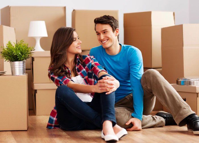 Couple sitting on the floor smiling at each other in a room with packed moving boxes.