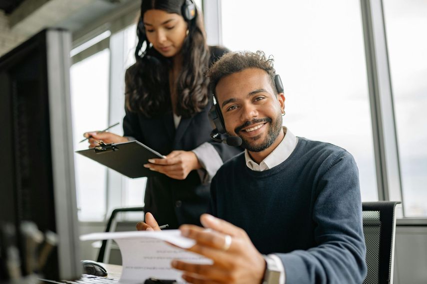 A man is sitting at a desk with a clipboard and a woman is standing behind him.
