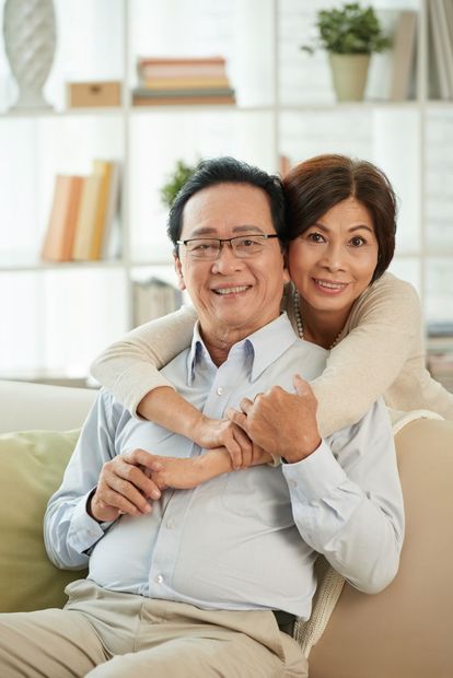 Smiling Asian couple embraces on a couch in a brightly lit living room.